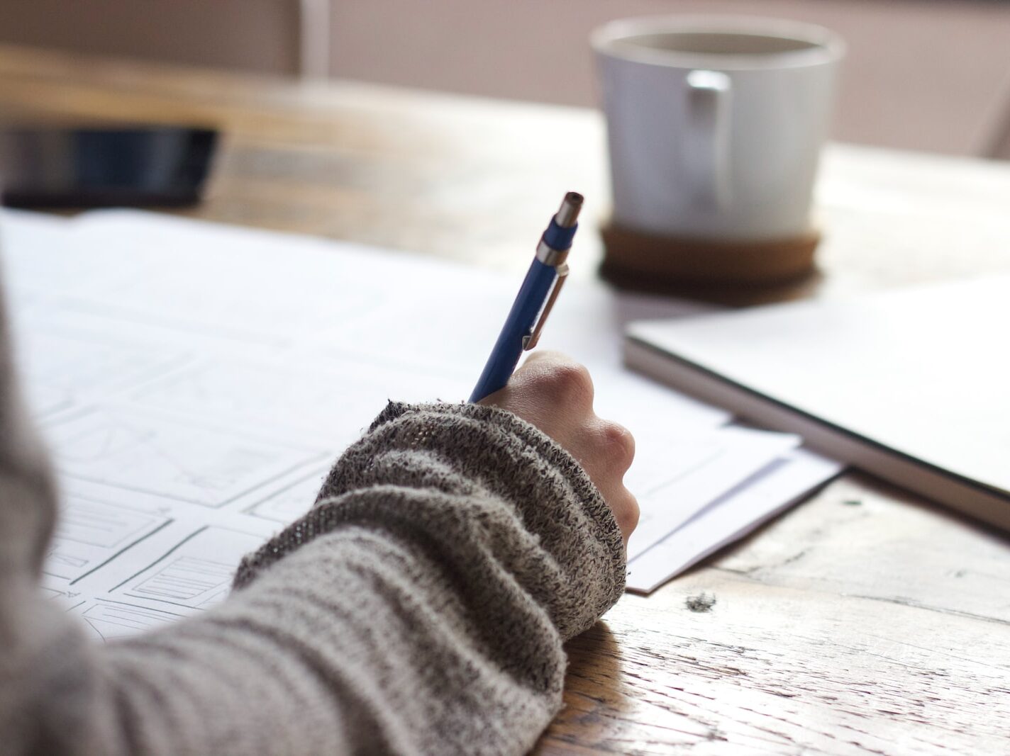 person writing on brown wooden table near white ceramic mug